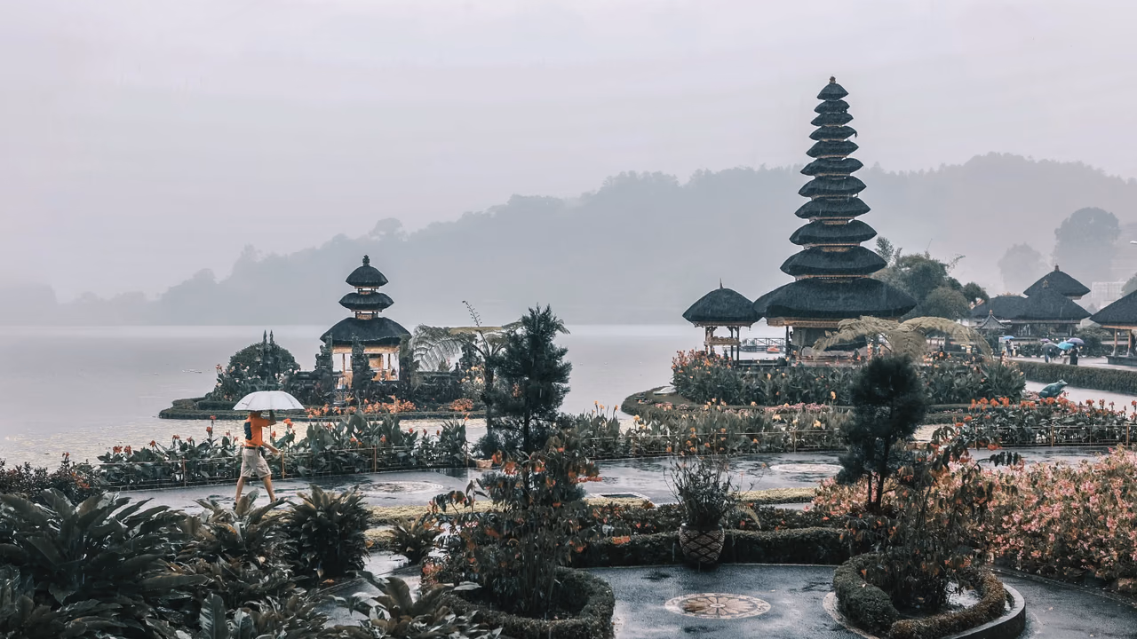 Traveler carrying an umbrella beside a lakeside temple in rainy weather