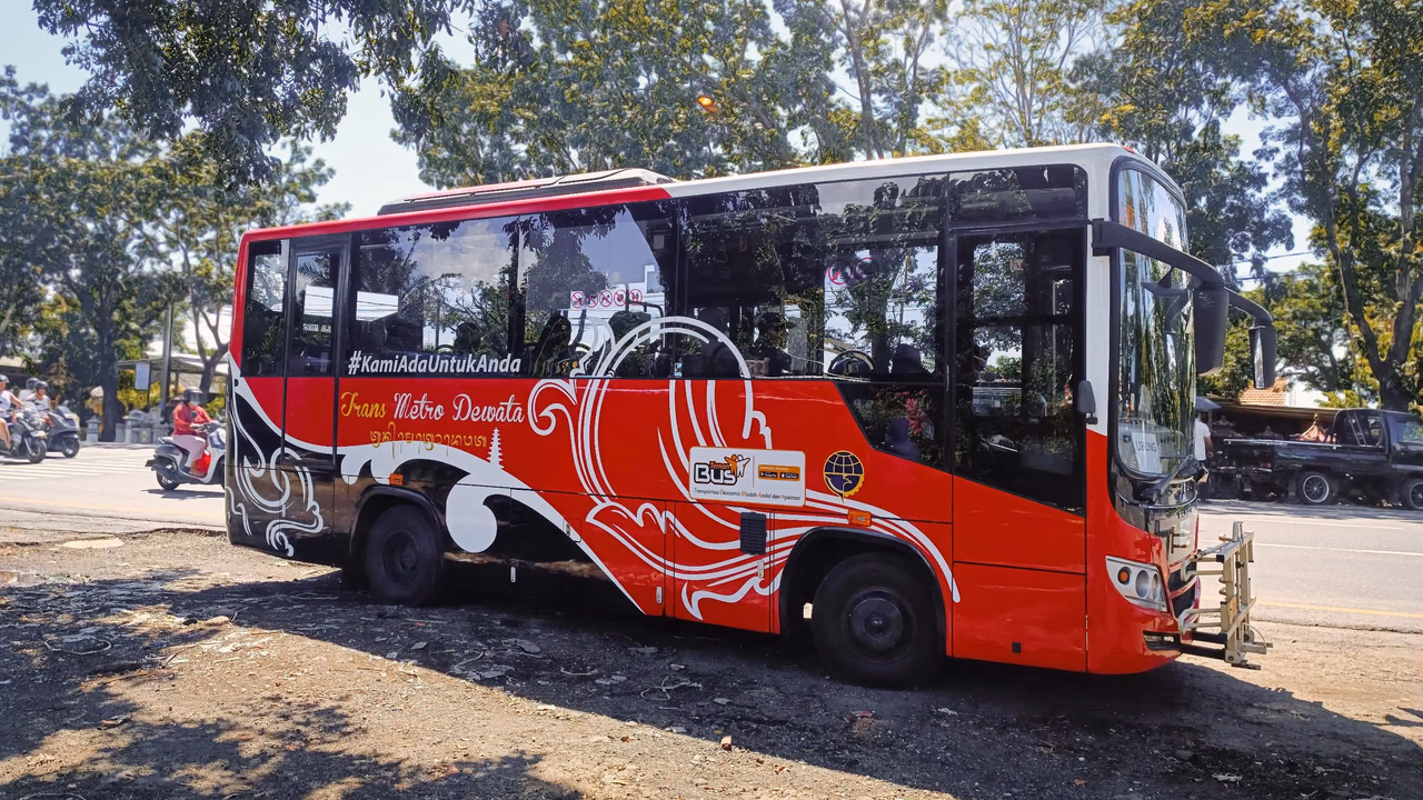 Red and white Trans Metro Dewata bus parked beside a tree-lined road in Bali